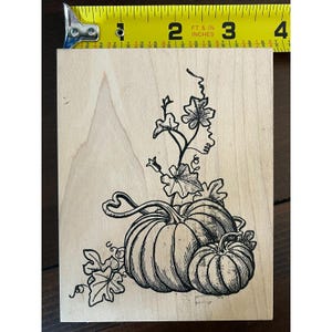 May include: A rectangular wooden rubber stamp featuring a black ink illustration of two pumpkins with vines and leaves. A yellow measuring tape is positioned at the top, showing the stamp width is approximately 3.5 inches across the wood block.