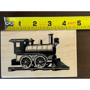 May include: A rectangular wooden rubber stamp featuring a black ink illustration of a steam locomotive train on tracks. The train has the word DART printed on the side and the number 1887 near the front. A yellow measuring tape shows it is 5 inches wide.