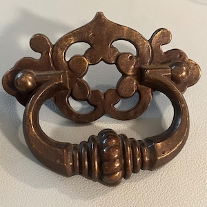 May include: Bronze-toned cabinet pull with an ornate, decorative backplate and a rounded handle. The backplate features a floral design with openwork details. The handle has a ribbed texture. This hardware is suitable for furniture restoration or new builds.
