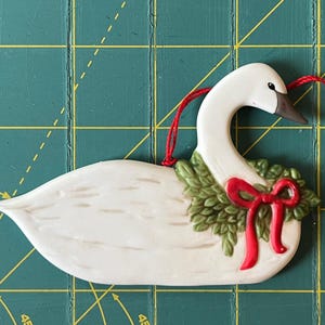 May include: A white ceramic swan ornament featuring a green wreath around its neck adorned with a red bow. The swan has a dark brown beak and a black eye. A thin red string is attached to the top of the swan for hanging purposes.