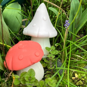 May include: Two ceramic mushroom figurines, one white with pink polka dots and one red with white polka dots, are nestled in a bed of green grass and small purple flowers.