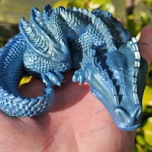 May include: A blue dragon figurine with a metallic finish. The dragon is curled up, with its head and tail visible. The figurine has detailed scales and a textured surface. The dragon is a fantasy-themed item.