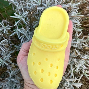 May include: A vibrant yellow clog shoe, with the brand name "Crocs" embossed on the side. The shoe has several ventilation holes and is held in a hand against a backdrop of green and grey foliage.
