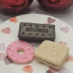 May include: A plate featuring a pink iced donut, a dark brown "Bourbon" biscuit, and a light-coloured biscuit with a decorative pattern. The plate has heart-shaped designs.