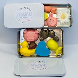May include: A white tin with a label that reads "Bubbles in a Tin Mini Shower Soap by Ladybirdsrco". The tin contains a variety of small, colourful soaps shaped like animals, hearts, and a pineapple. The soaps are arranged in two rows.