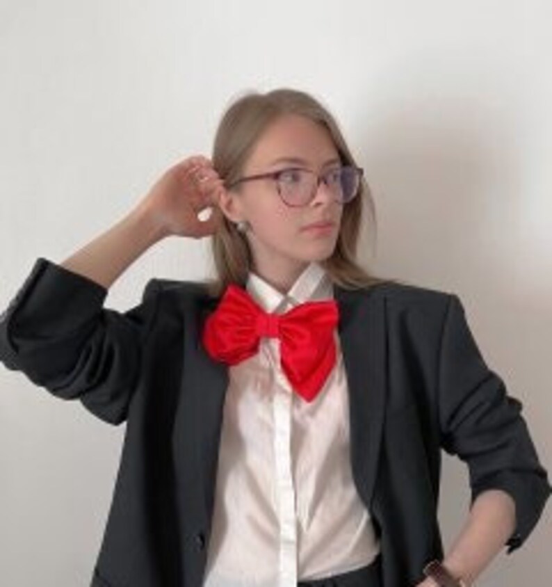 May include: A woman wearing a black blazer, a white collared shirt, and a large red bow tie.