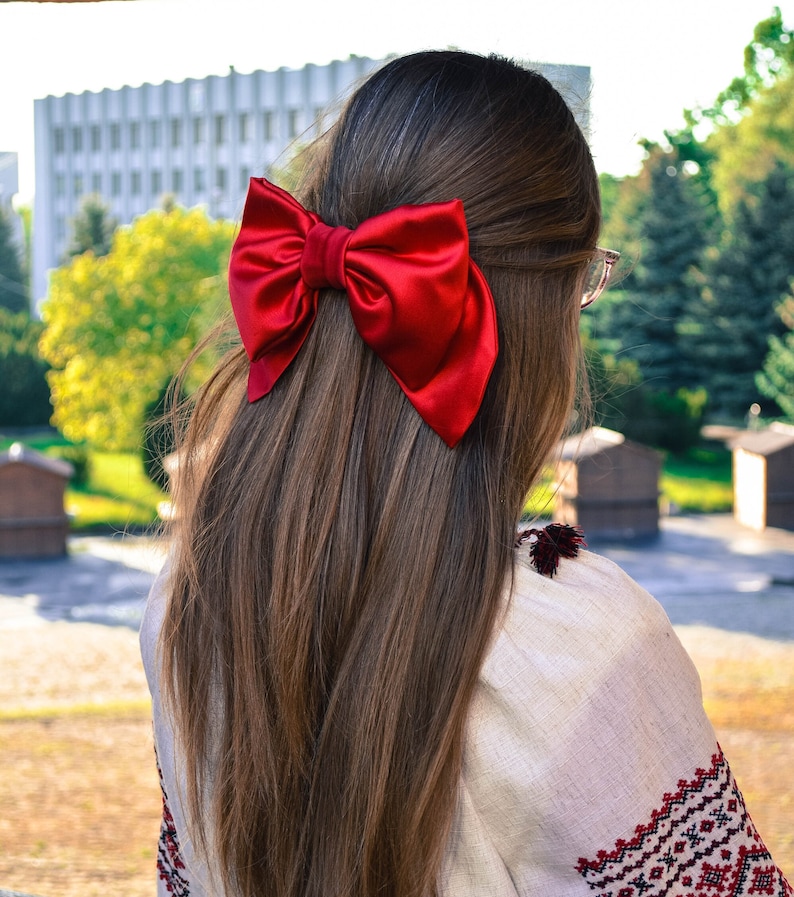 May include: A woman with long brown hair wearing a white embroidered blouse with a large red satin bow in her hair.