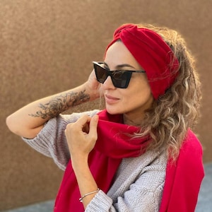 Op de afbeelding: Een vrouw draagt een rode turban hoofdband en een rode sjaal. Ze draagt een zwarte zonnebril en een grijze trui.