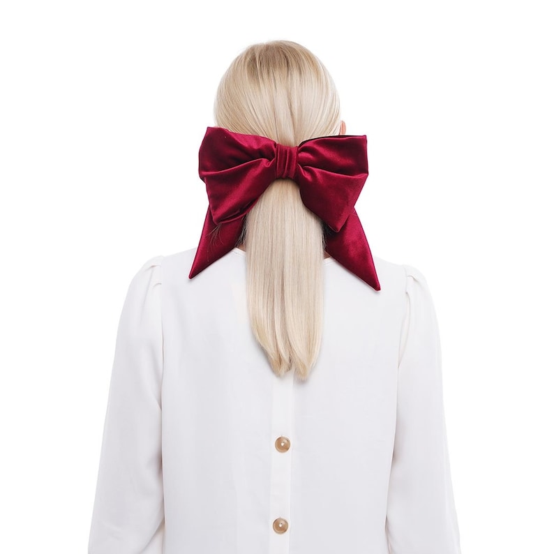 May include: A woman with blonde hair wearing a white blouse with buttons down the back. She is wearing a large, red velvet bow in her hair.