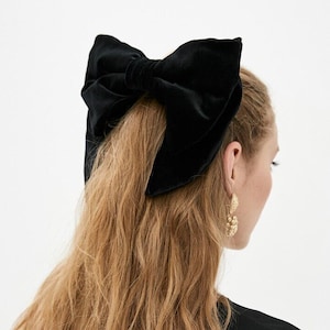 May include: A large black velvet bow hair clip. The bow is tied in a large, dramatic knot and is worn in the hair of a person with long, blonde hair.