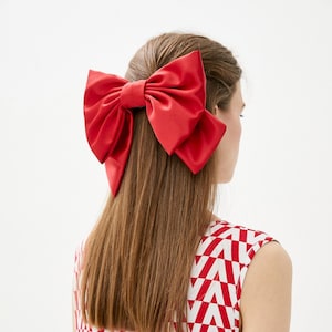 May include: A red satin bow hair clip. The bow is large and has a center loop. The hair clip is worn in a woman's long brown hair.