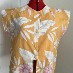 Op de afbeelding: Een mouwloze mosterdgele top met een botanische print van witte en roze bladeren en bloemen. Het kledingstuk heeft een ronde halslijn, een knoopsluiting aan de voorzijde met twee gele knopen en een textuur met een subtiel patroon.