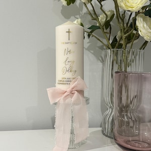 May include: A cream baptism candle with gold script and a cross. It features a light pink ribbon and sits on a clear glass base. White flowers and vases are in the background, creating an elegant display.