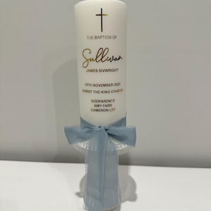 May include: A white baptism candle with gold lettering and a cross. The text reads "THE BAPTISM OF Sullivan JAMES SIVWRIGHT 15TH NOVEMBER 2025 CHRIST THE KING CHURCH GODPARENTS AMY FARR CAMERON LEY." A light blue ribbon and lace are tied around the base.