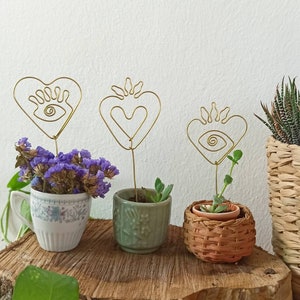 Puede incluir: Tres pequeñas plantas en maceta con esculturas de alambre dorado en forma de corazones y ojos. Las plantas están en macetas de diferentes colores y están sobre una superficie de madera.