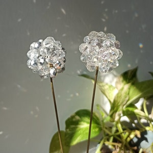 May include: Two crystal flower sculptures with clear glass beads on thin gold stems. The sculptures are in front of a green leafy plant in a pot.