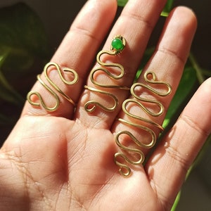 以下が含まれることがあります： 中指に緑色の宝石が付いた、金色のワイヤー製のリングが3つ。リングは波打つような蛇のようなパターンでデザインされています。