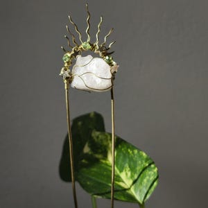 以下が含まれることがあります： 白いクリスタルストーンをあしらった手作りのヘアアクセサリー。金色のワイヤーと緑色のビーズで装飾されています。デザインはサンバーストのようなクラウンを含みます。アクセサリーは2本の長い金色のワイヤーで保持されています。