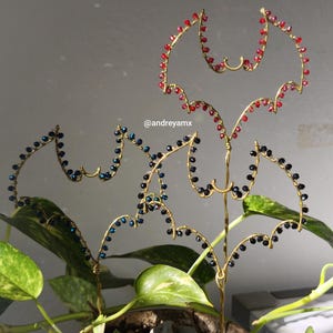 May include: Gold wire bat-shaped plant stakes adorned with red and blue beads. The stakes are placed in a potted plant with green and yellow leaves. The @andreyamx watermark is visible.