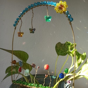 May include: A wire sculpture of a rainbow arch with hanging butterflies and a yellow flower at the top. The sculpture is placed in a pot with a green plant and colorful beads.