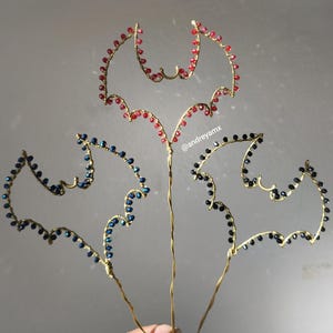 May include: Three handmade bat-shaped wire ornaments. Each bat is crafted with gold-colored wire and adorned with colorful beads. One bat features red beads, another blue, and the third black. The ornaments are held in a hand.