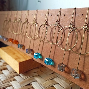 May include: A display of handmade earrings with double hoop designs and colorful crystal accents. The earrings are displayed on a wooden board with a woven background.