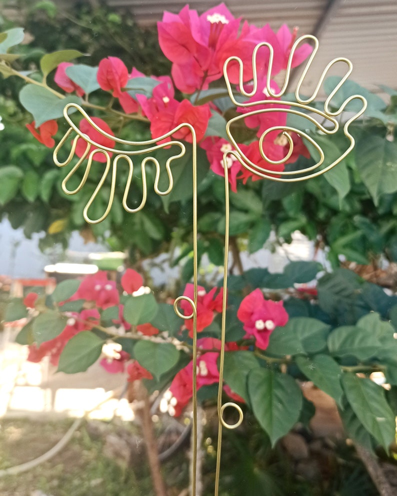 May include: Gold wire sculpture of an eye and eyelashes against a background of pink bougainvillea flowers.