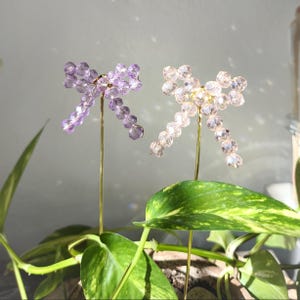 May include: Two decorative plant stakes with bow-shaped designs. One bow is made of lavender-colored beads, and the other is made of clear beads. Both bows are attached to thin, gold-colored stems. The stakes are placed in a pot with green plants.