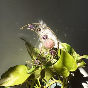 May include: A gold wire crescent moon with a pink and white gemstone in the center. The moon is decorated with smaller purple and clear gemstones.