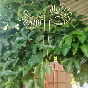 May include: Gold wire sculpture of a stylized eye with eyelashes. The sculpture is made of thin wire and has a simple, modern design.