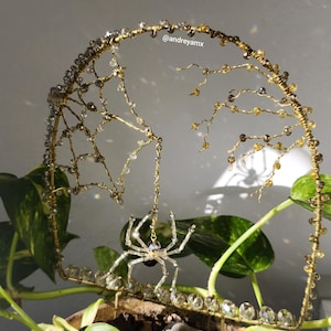 May include: A decorative gold-coloured wire sculpture featuring a spider and web design, embellished with clear and amber beads. The spider hangs from the web, with the artist's handle, @andreyamx, visible. The sculpture is set against a backdrop of green foliage.