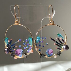 May include: Gold hoop earrings with a whimsical design featuring iridescent blue, purple, and green beads, black stars, and a pink shell.