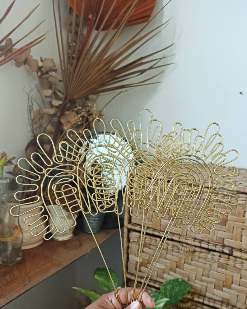 Pu&ograve; includere: Set di tre sculture in filo dorato a forma di archi con un design ondulato decorativo. Le sculture sono disposte a ventaglio e sono tenute nella mano di una persona.