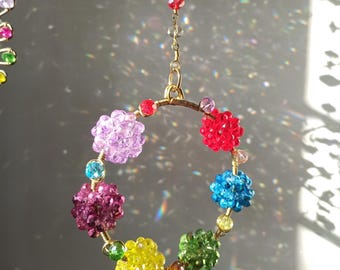 May include: A vibrant, beaded suncatcher with a gold-coloured metal ring and chain. It features clusters of faceted beads in various colours, including red, blue, green, yellow, and purple. The beads are arranged in a circular pattern.