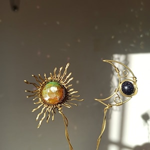 Può includere: Un paio di fermagli per capelli in filo d'oro con accenti di pietre preziose. Un fermaglio ha la forma di un sole con una pietra di agata multicolore, e l'altro ha la forma di una mezzaluna con una pietra blu scuro.