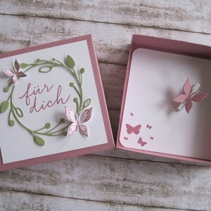 May include: Open gift box with a white card featuring a green wreath and pink butterflies. The card reads "f&uuml;r dich" in pink script. The box is pink and contains a card with pink butterfly accents.