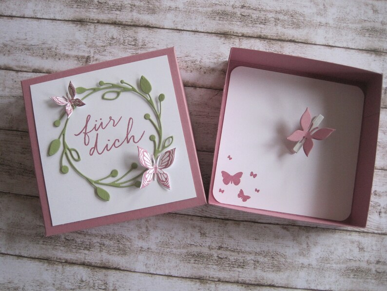 May include: Open gift box with a white card featuring a green wreath and pink butterflies. The card reads "f&uuml;r dich" in pink script. The box is pink and contains a card with pink butterfly accents.