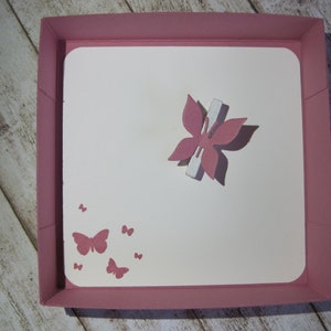 May include: A pink square box with a white interior. A pink butterfly with a white accent is in the center. Smaller pink butterflies are printed in the lower left corner. The box is on a light wood surface.