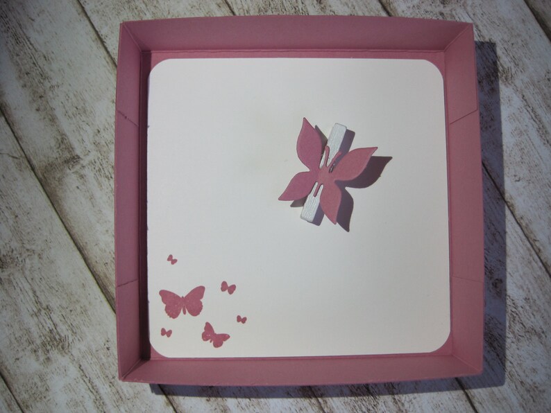 May include: A pink square box with a white interior. A pink butterfly with a white accent is in the center. Smaller pink butterflies are printed in the lower left corner. The box is on a light wood surface.