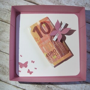 May include: A pink square gift box with a white interior holds a ten Euro bill and a pink flower embellishment. Pink butterfly stamps decorate the white interior. The box sits on a light wood-grain surface.