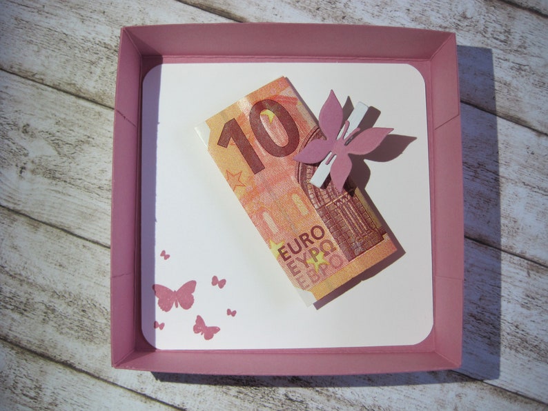 May include: A pink square gift box with a white interior holds a ten Euro bill and a pink flower embellishment. Pink butterfly stamps decorate the white interior. The box sits on a light wood-grain surface.