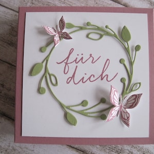 May include: A square greeting card with a white center and a mauve border. The card features a green wreath with leaves and two pink butterflies. The words "f&uuml;r dich" are written in mauve script within the wreath.