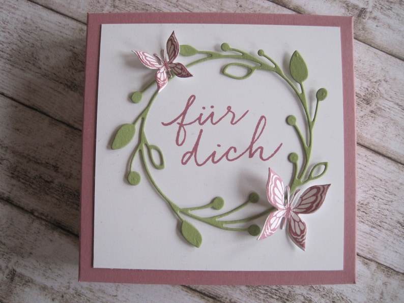May include: A square greeting card with a white center and a mauve border. The card features a green wreath with leaves and two pink butterflies. The words "f&uuml;r dich" are written in mauve script within the wreath.