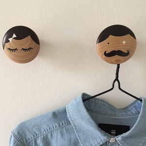 Könnte beinhalten: Zwei hölzerne Wandhaken in Form von Gesichtern. Der weibliche Haken hat geschlossene Augen und eine schwarze Schleife. Der männliche Haken hat einen Schnurrbart und ein schwarzes Pluszeichen auf seinem Gesicht. Ein blaues Jeanshemd hängt am männlichen Haken.