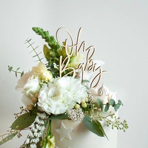 May include: White cake with a wooden "Oh Baby" topper and white flowers. The cake is decorated with white stars.