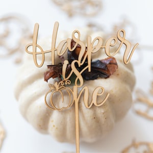 May include: Wooden cake topper with the text "Harper is one" and a pumpkin design. The topper is on a white pumpkin.
