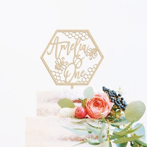 May include: A wooden cake topper in the shape of a hexagon with a honeycomb pattern and the text "Amelia is One" with bees. The topper is on a white cake with pink and green flowers.
