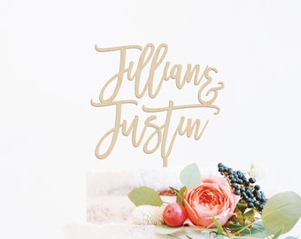 Puede incluir: Topper de pastel de madera con los nombres "Jillian & Justin" escritos en cursiva. El topper está colocado sobre un pastel blanco decorado con vegetación y flores rosas.