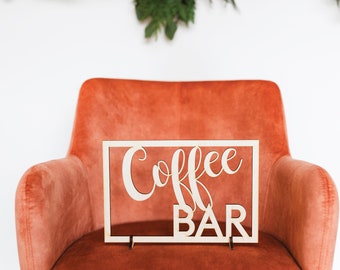 Könnte beinhalten: Ein Holzschild mit den Worten "Coffee Bar" in einer kursiv geschriebenen Schrift, das auf einem orangefarbenen Samtstuhl steht.