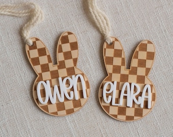 May include: Two wooden bunny-shaped ornaments with checkered patterns and white names "Owen" and "Clara" on them. The ornaments are attached to white strings.
